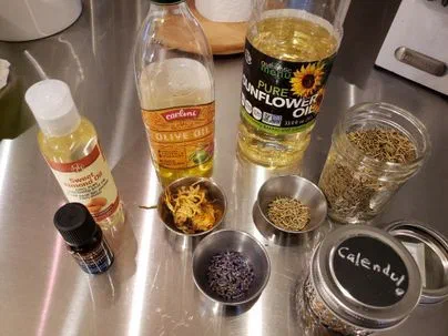 Sweet almond, olive and sunflower oil on a steel counter beside small bowls of dried calendula and lavender and a jar of dried herb labelled Calendula
