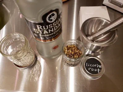 A bottle of vodka on a steel counter with an empty jar, a small jar of crushed root, and a lid labelled licorice root