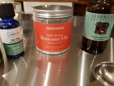 A jar of Indian Healing bentonite clay between a bottle of tea tree essential oil and a bottle of witch hazel extract, on a steel counter