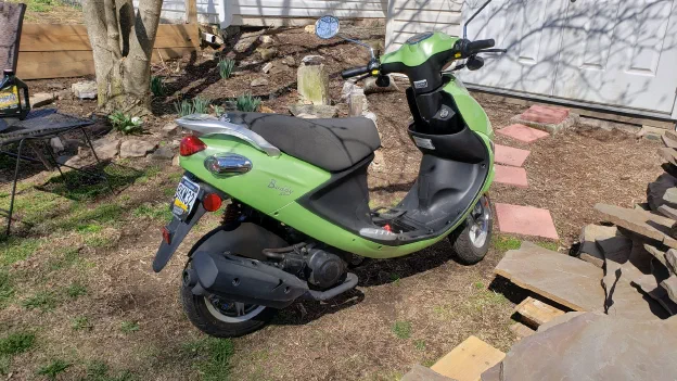 A green motor scooter parked beside a garden bed in early spring