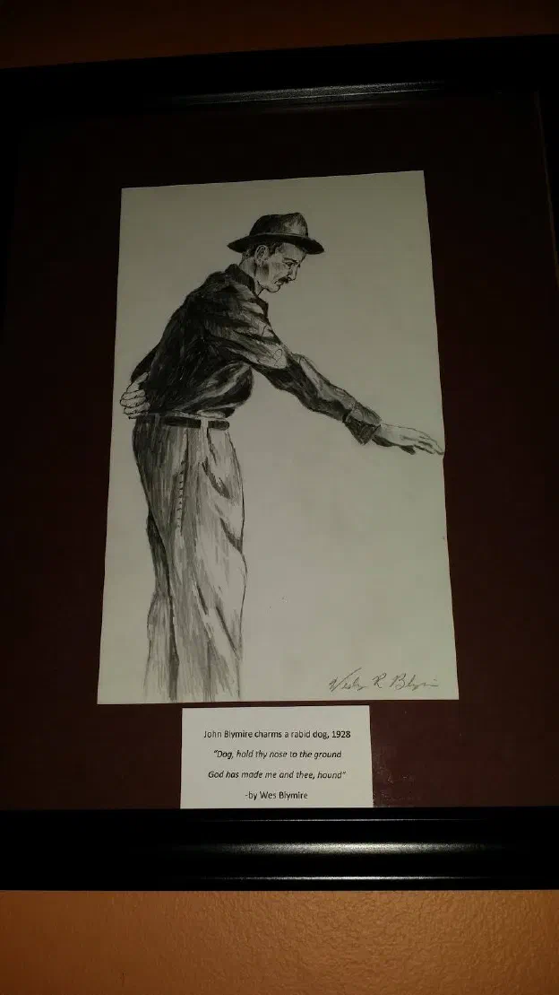 A framed pencil drawing of John Blymire bending toward a dog with one hand outstretched, signed Wes R. Blymire, a printed plaque beneath it