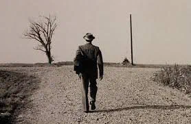 A lone figure in a hat walking away down a dirt road in open country