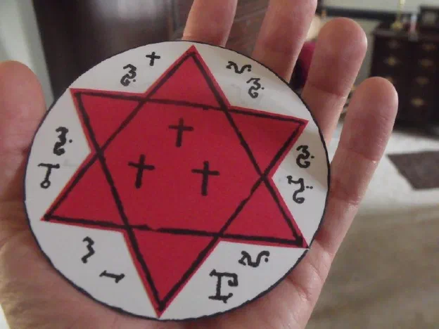A finished paper talisman held in the palm — a red six-pointed star with three crosses on a white disc, planetary sigils around the rim