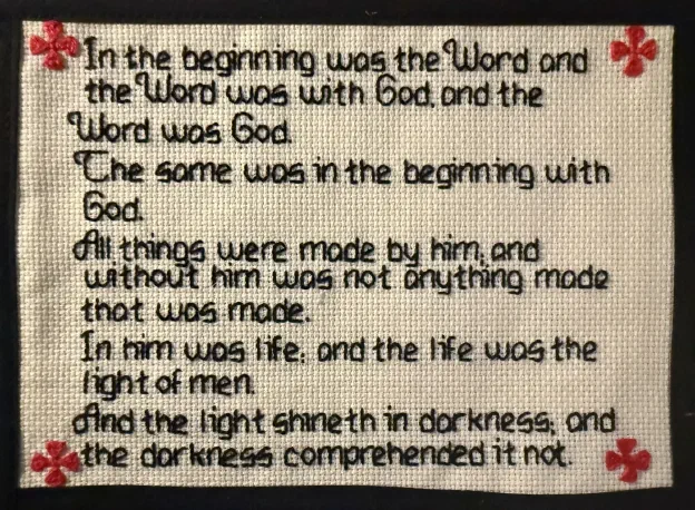 A cross-stitched panel on cream Aida cloth, mounted on black. In black thread, the English text of John 1:1–5, beginning In the beginning was the Word and ending and the darkness comprehended it not. A red cross is stitched in each of the four corners.